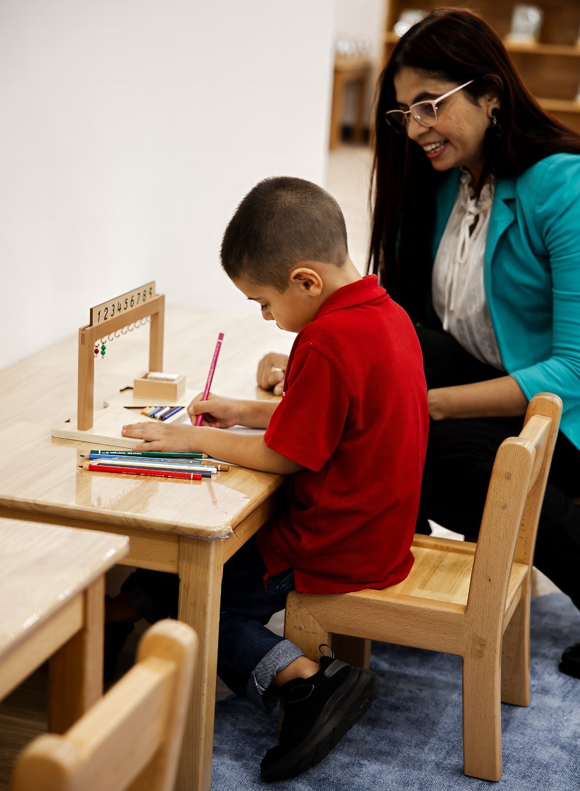 A child writing at a table with an educator nearby